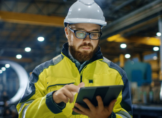 Industrial Engineer in Hard Hat Wearing Safety Jacket Uses Touchscreen Tablet Computer. He Works at the Heavy Industry Manufacturing Factory.