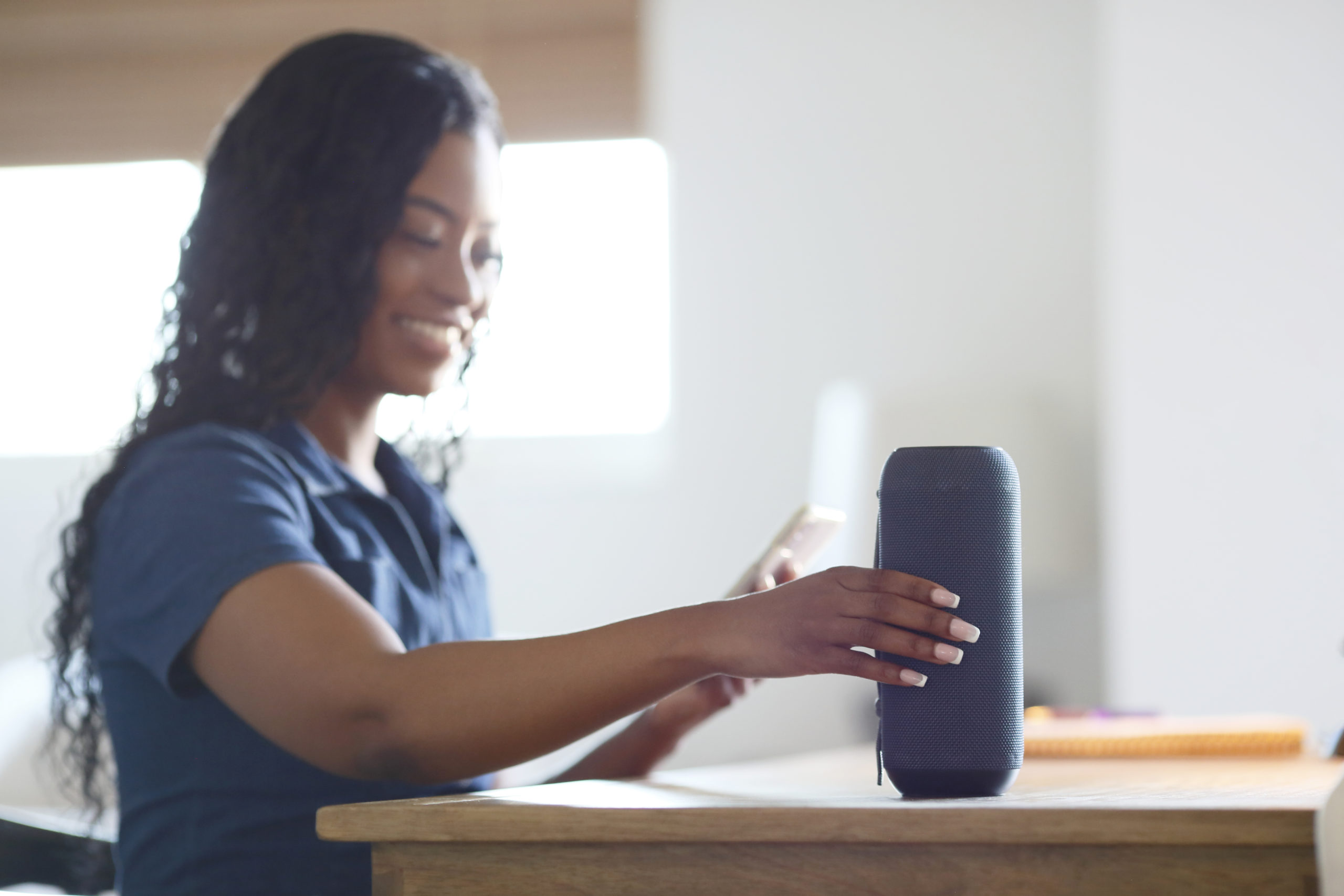 An African American millennial woman with long hair, sits at a wooden table while setting up a wireless speaker with her cell phone.